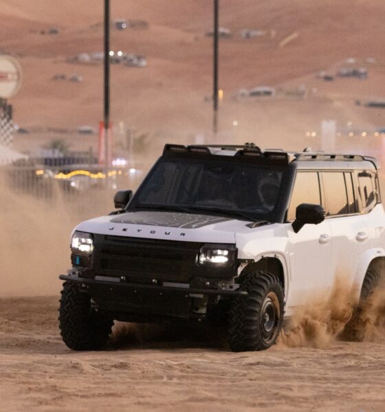 A white Jetour T2 off-road vehicle kicks up a large cloud of sand as it drives across the desert during the day. The vehicle is modified with a black hood decal and a roof rack. The background shows the hazy outlines of desert structures and festival signage under a sunset sky.