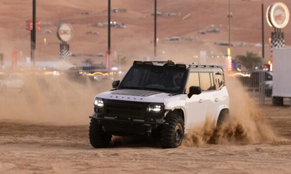 A white Jetour T2 off-road vehicle kicks up a large cloud of sand as it drives across the desert during the day. The vehicle is modified with a black hood decal and a roof rack. The background shows the hazy outlines of desert structures and festival signage under a sunset sky.