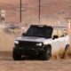 A white Jetour T2 off-road vehicle kicks up a large cloud of sand as it drives across the desert during the day. The vehicle is modified with a black hood decal and a roof rack. The background shows the hazy outlines of desert structures and festival signage under a sunset sky.