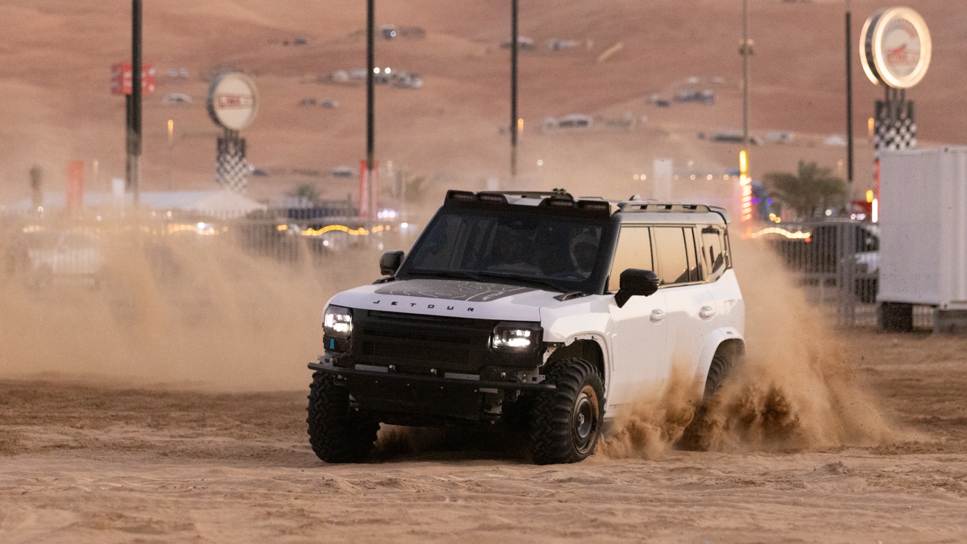 A white Jetour T2 off-road vehicle kicks up a large cloud of sand as it drives across the desert during the day. The vehicle is modified with a black hood decal and a roof rack. The background shows the hazy outlines of desert structures and festival signage under a sunset sky.