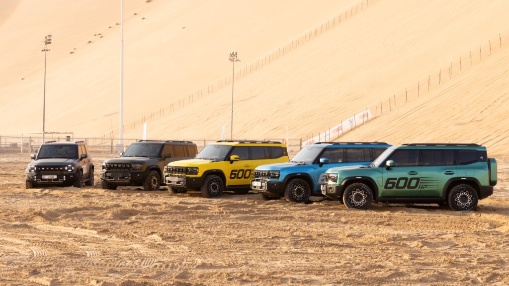 Five Jetour T2 SUVs in different colors (grey, black, yellow, blue, and teal) are parked in a row on the sand at the base of a massive dune. The teal and yellow vehicles prominently feature "600 HP" graphics on their side doors. The scene is bathed in the warm, soft light of late afternoon.