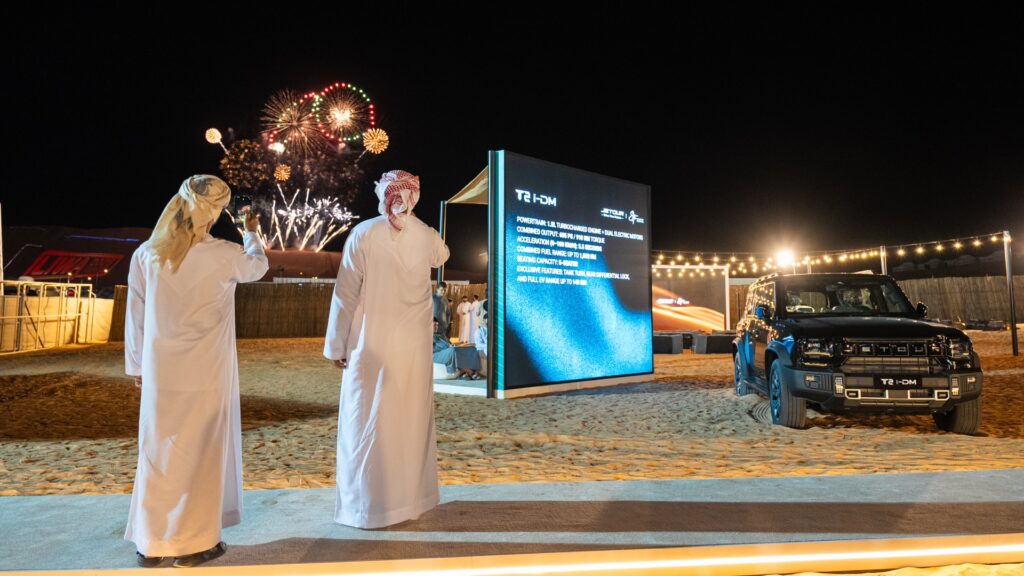 A wide night shot at the Liwa Festival featuring two men in traditional white Emirati kanduras watching a fireworks display. To the right, a black Jetour T2 i-DM is parked next to a large illuminated digital screen displaying technical specifications. The desert ground is sandy, and strings of warm decorative lights are visible in the background.