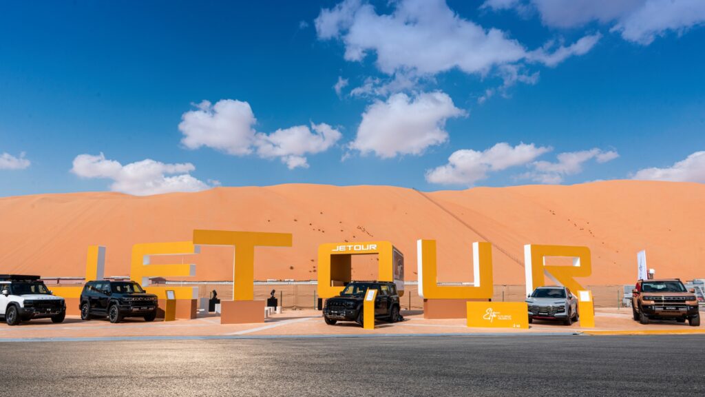 A daytime view of a desert exhibition featuring massive, standalone yellow and white letters spelling out "JETOUR" against a backdrop of a towering orange sand dune. Several Jetour SUVs in various colors—white, black, and silver—are parked in front of and between the letters. The sky is bright blue with scattered white clouds.
