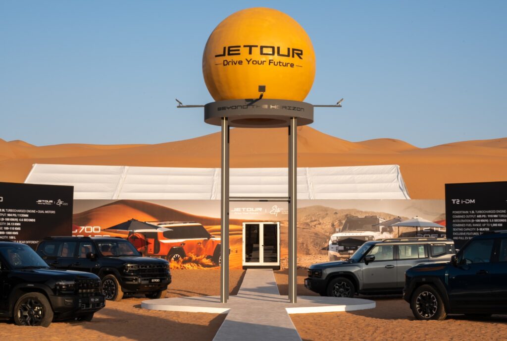 A large yellow spherical monument with the "JETOUR - Drive Your Future" logo sits atop a tall metal structure in the desert. Below it, a white walkway leads toward a small building. Several black and grey Jetour T2 i-DM SUVs are parked on the surrounding sand, with a massive sand dune and white festival tents in the background.