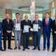 Group of executives standing indoors at Al Habtoor Grand Resort, holding framed ISO certification documents, with a modern decorative pillar in the background.