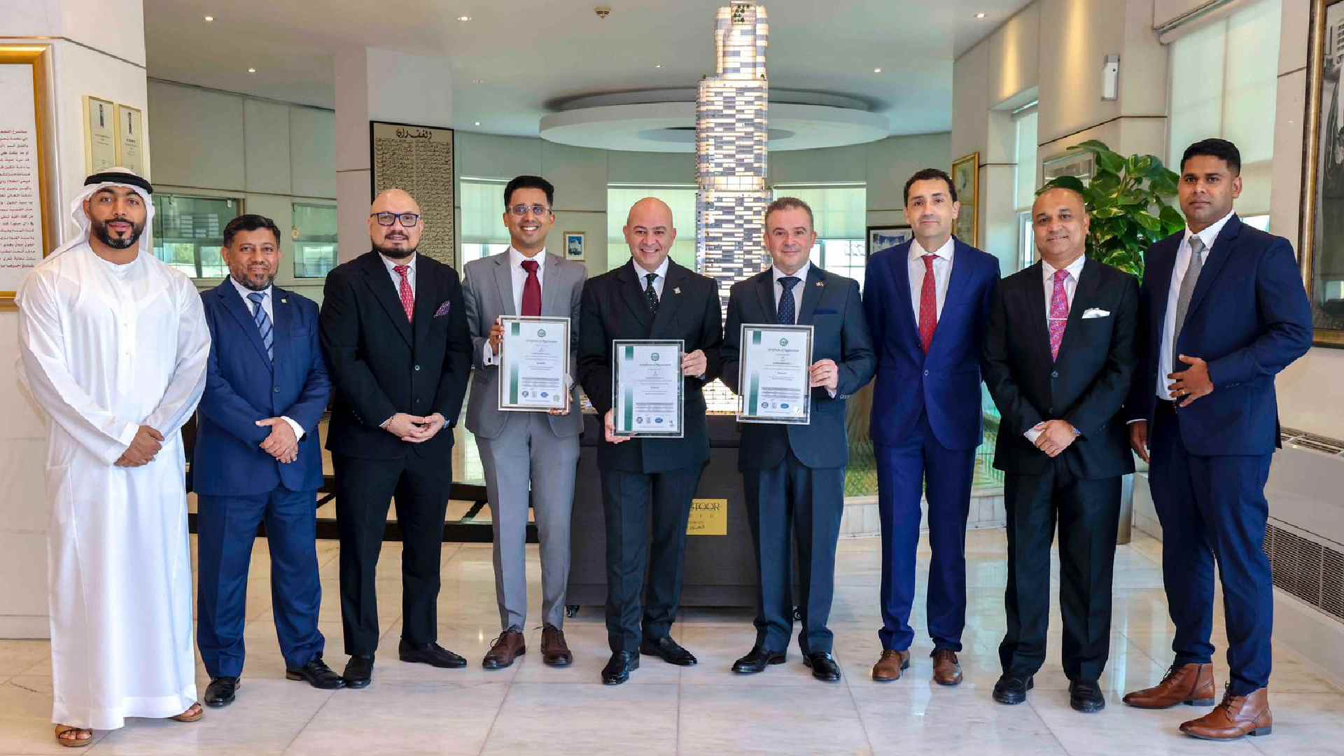 Group of executives standing indoors at Al Habtoor Grand Resort, holding framed ISO certification documents, with a modern decorative pillar in the background.