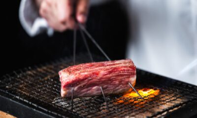 Grilled Japanese Wagyu beef being cooked over an open flame on a wire mesh grill at KEN BY KAMATSUDA, held with metal skewers for even roasting.