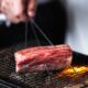 Grilled Japanese Wagyu beef being cooked over an open flame on a wire mesh grill at KEN BY KAMATSUDA, held with metal skewers for even roasting.