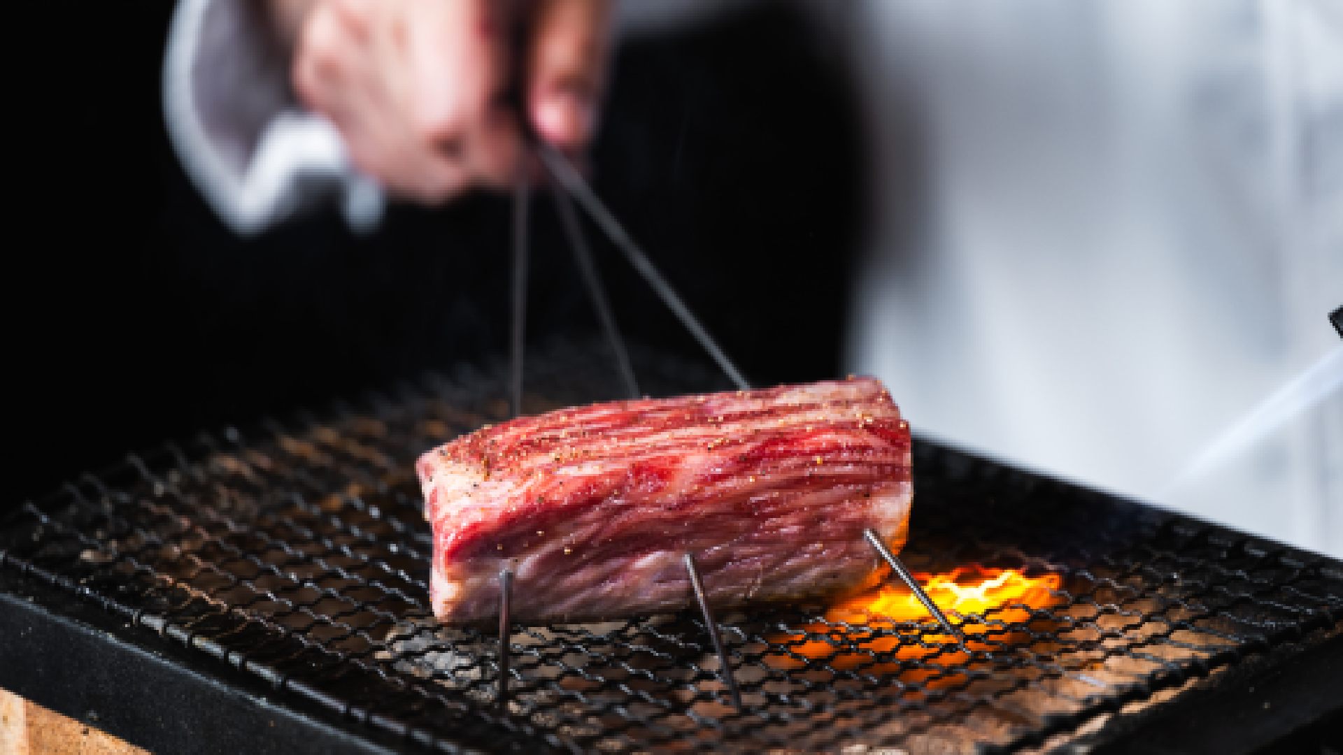 Grilled Japanese Wagyu beef being cooked over an open flame on a wire mesh grill at KEN BY KAMATSUDA, held with metal skewers for even roasting.