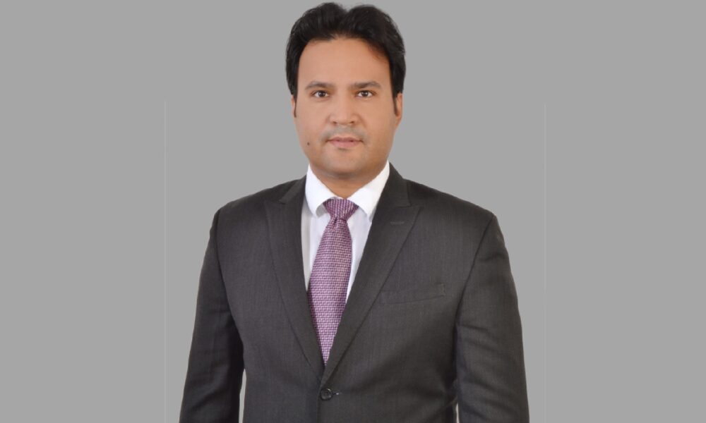 Person wearing a dark gray business suit with a white dress shirt and a textured purple tie, standing against a plain gray background