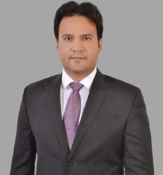 Person wearing a dark gray business suit with a white dress shirt and a textured purple tie, standing against a plain gray background