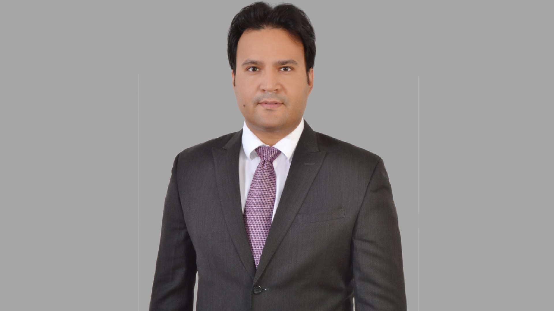 Person wearing a dark gray business suit with a white dress shirt and a textured purple tie, standing against a plain gray background