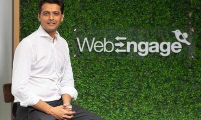 Person seated in front of a green artificial foliage wall featuring the WebEngage logo with a bird icon.