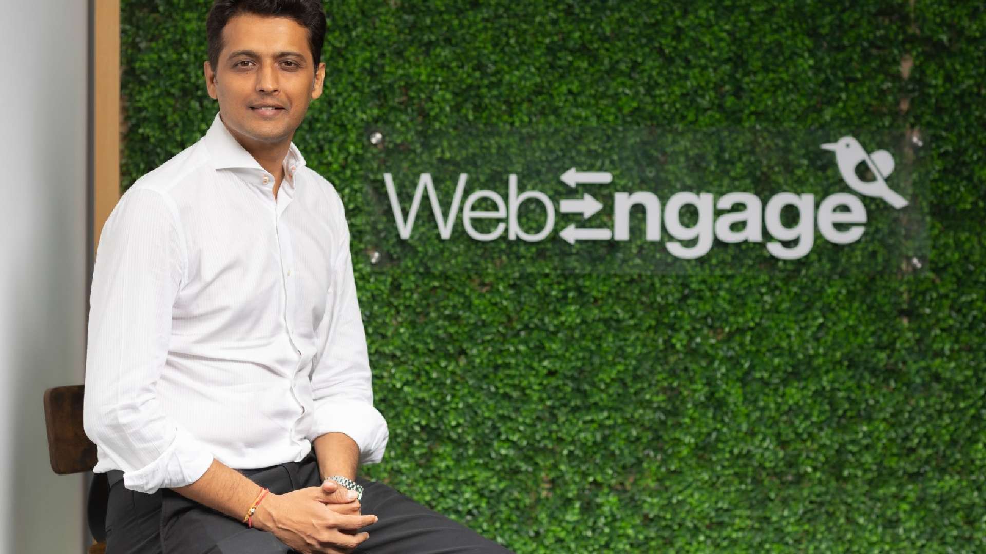 Person seated in front of a green artificial foliage wall featuring the WebEngage logo with a bird icon.