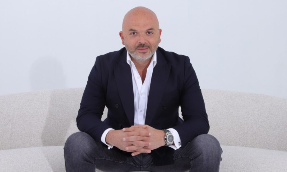 A person wearing a dark blazer over a white dress shirt and jeans, seated on a light-colored sofa with hands clasped together, showcasing a large wristwatch.