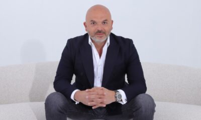 A person wearing a dark blazer over a white dress shirt and jeans, seated on a light-colored sofa with hands clasped together, showcasing a large wristwatch.