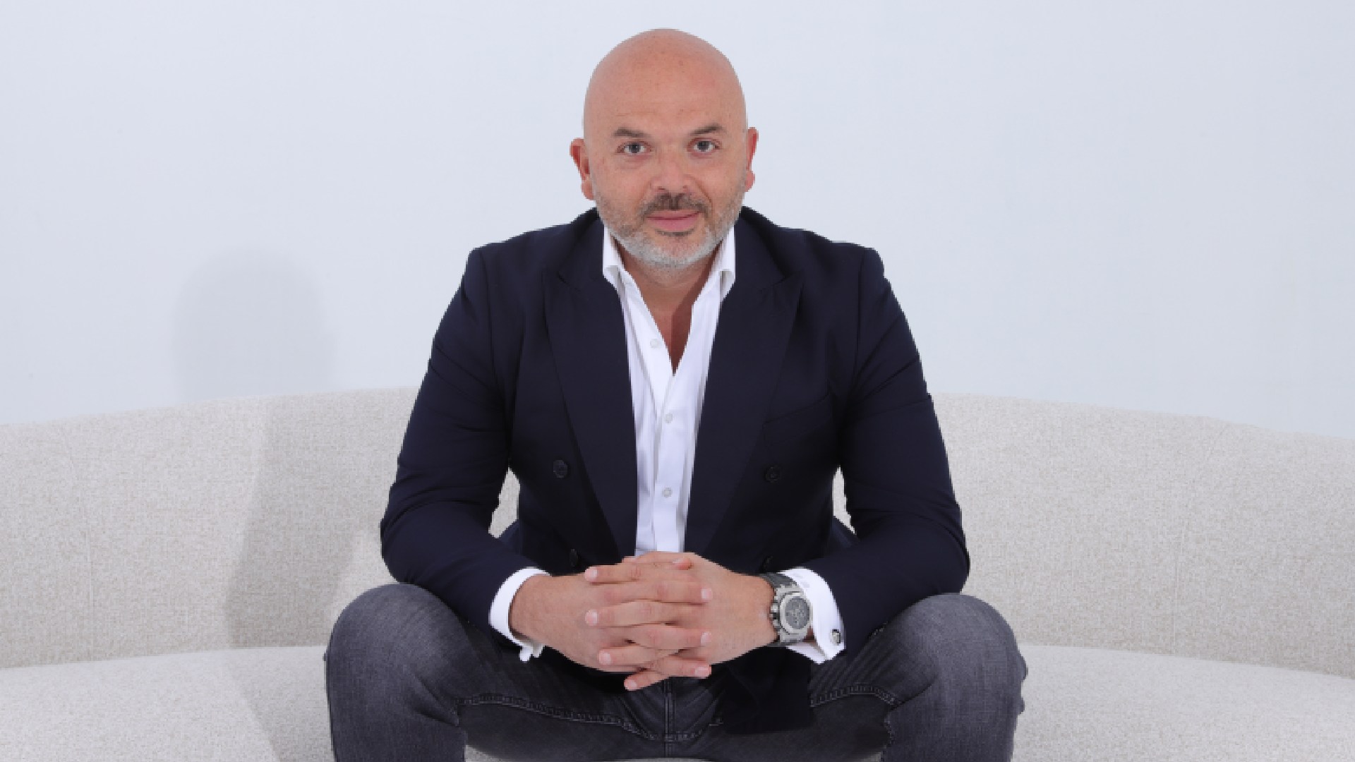 A person wearing a dark blazer over a white dress shirt and jeans, seated on a light-colored sofa with hands clasped together, showcasing a large wristwatch.