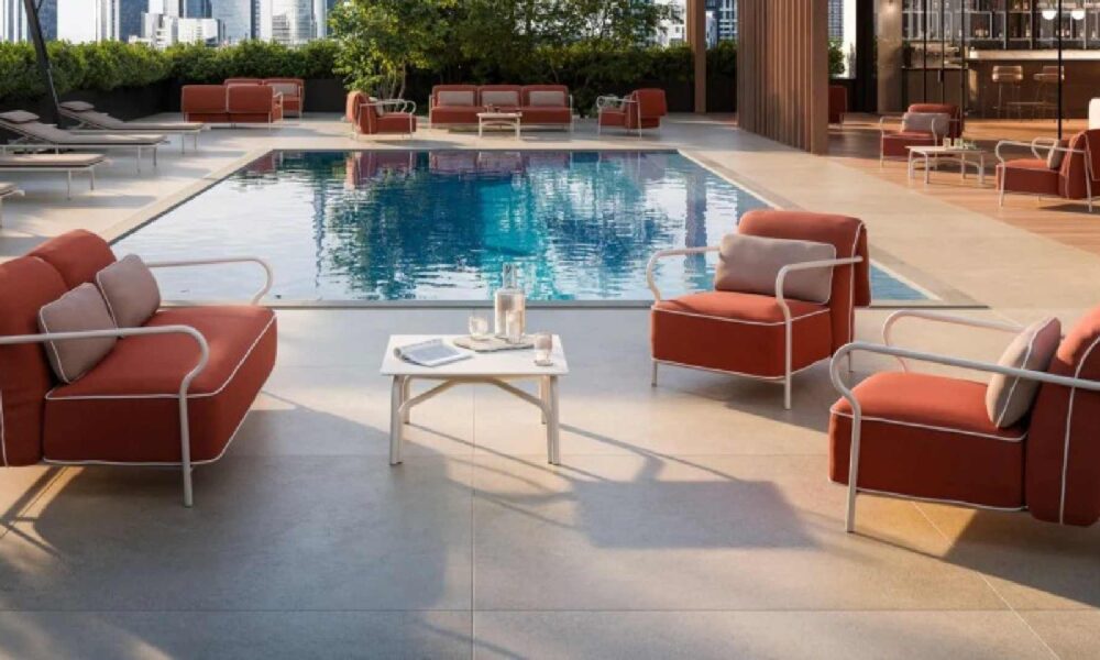 Outdoor poolside lounge area featuring modern red cushioned armchairs and a matching sofa set with white metal frames, arranged around a small white coffee table holding a glass pitcher and two glasses. The setting overlooks a clear blue swimming pool, surrounded by additional seating and greenery in the background.