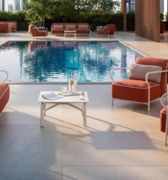 Outdoor poolside lounge area featuring modern red cushioned armchairs and a matching sofa set with white metal frames, arranged around a small white coffee table holding a glass pitcher and two glasses. The setting overlooks a clear blue swimming pool, surrounded by additional seating and greenery in the background.