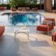 Outdoor poolside lounge area featuring modern red cushioned armchairs and a matching sofa set with white metal frames, arranged around a small white coffee table holding a glass pitcher and two glasses. The setting overlooks a clear blue swimming pool, surrounded by additional seating and greenery in the background.