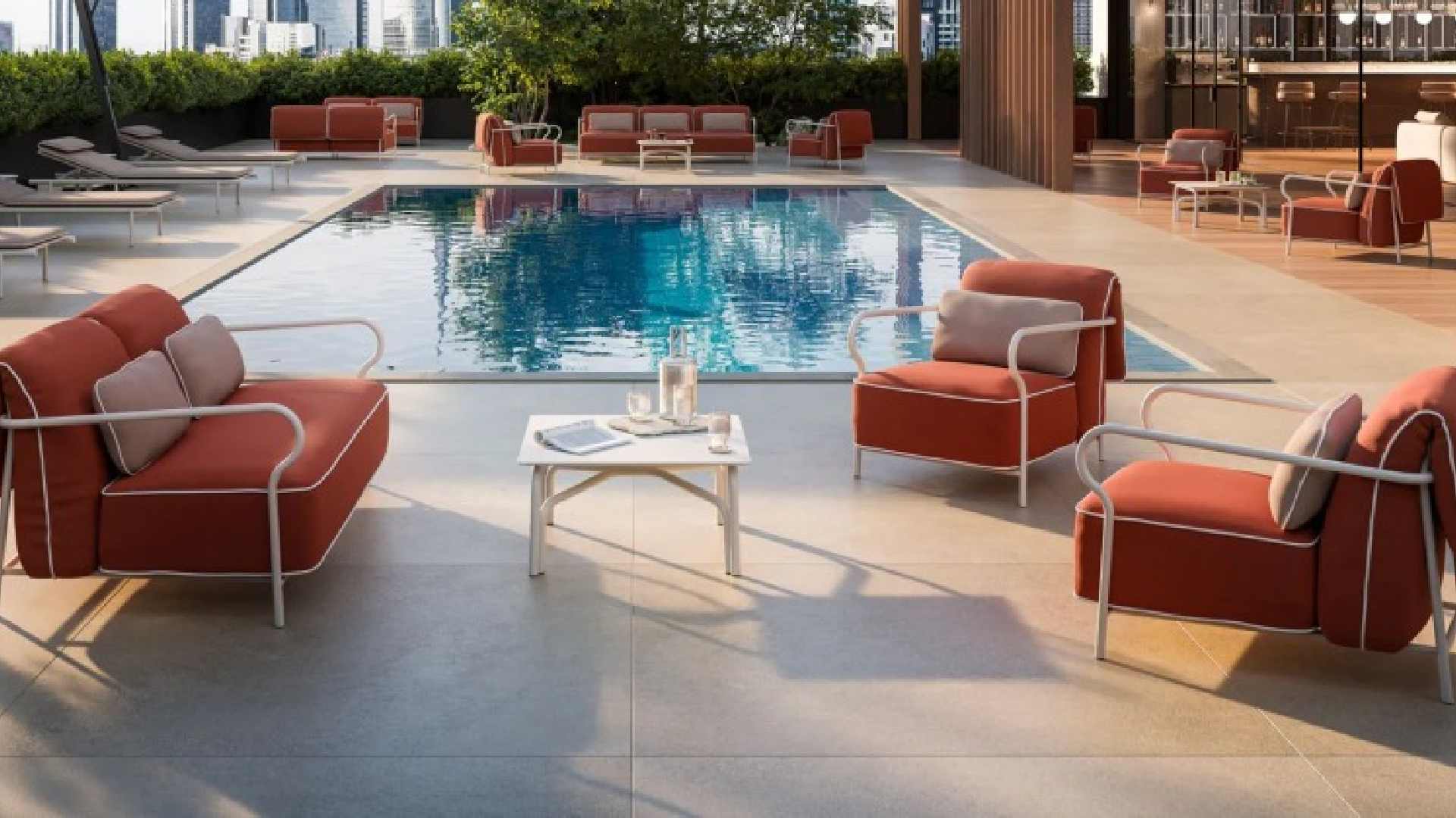 Outdoor poolside lounge area featuring modern red cushioned armchairs and a matching sofa set with white metal frames, arranged around a small white coffee table holding a glass pitcher and two glasses. The setting overlooks a clear blue swimming pool, surrounded by additional seating and greenery in the background.