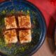 Four square pieces of Turkish dessert served on a dark blue ceramic plate, garnished with ground pistachios and cinnamon powder, placed on a wooden table with a red decorative element in the background.