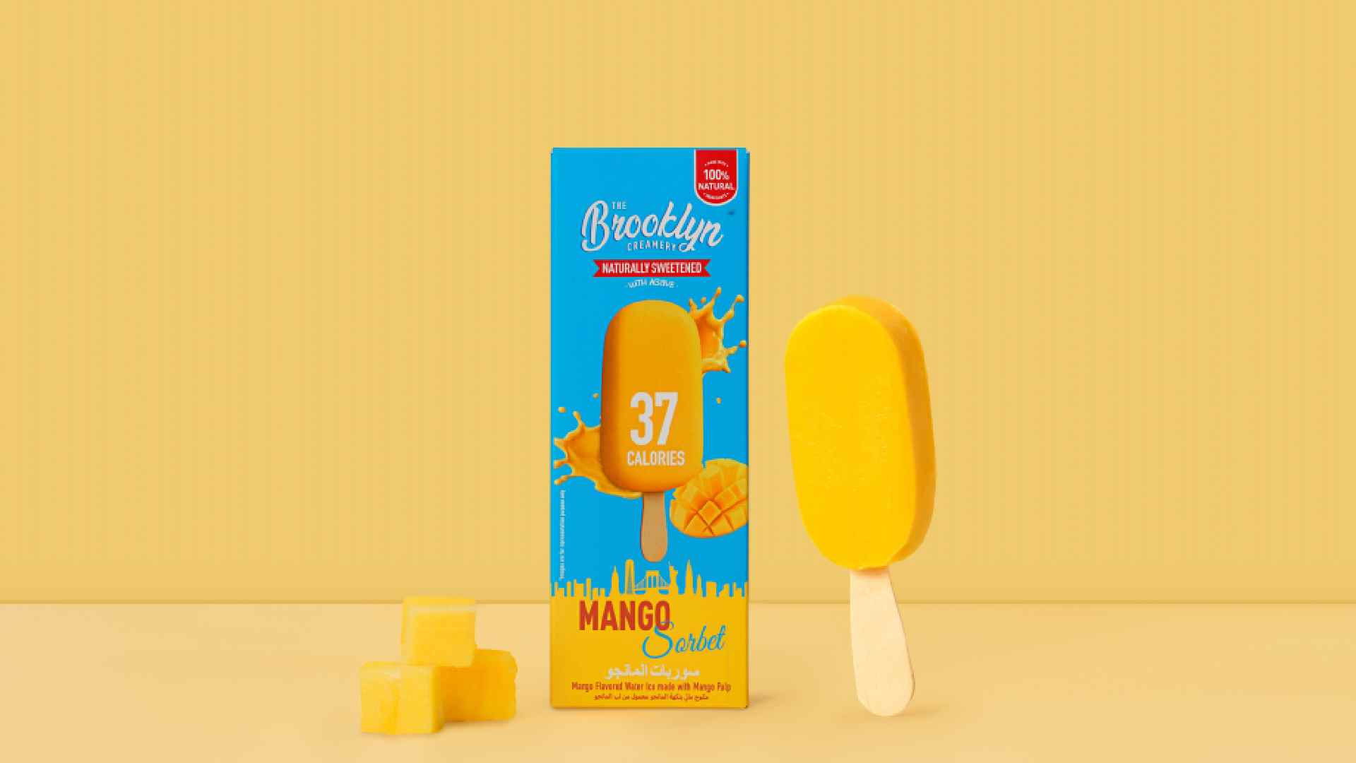 Mango sorbet bar from The Brooklyn Creamery displayed next to its bright blue and orange packaging that reads ‘37 Calories’ and ‘Mango Sorbet.’ The box features splashes of mango juice and a mango graphic, with three small mango cubes placed in front on a yellow background.