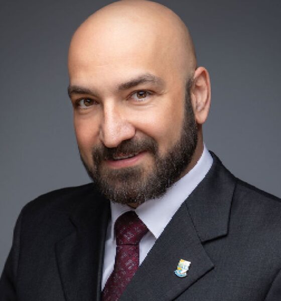 A professional man with short dark hair and a suit jacket is giving a presentation to a diverse group of seated individuals in a formal conference room with a large screen displaying charts behind him.