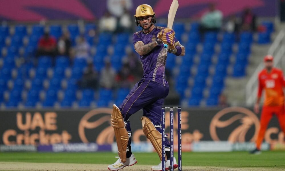A professional cricket player for the Desert Vipers in mid-swing during a match. The batsman is wearing a dark green and black patterned jersey with red accents, a red helmet, and black protective leg pads. He is holding a wooden cricket bat high in a follow-through motion after playing a shot. The background shows a crowded stadium with purple and blue seating and a "DP World" branded wicket.
