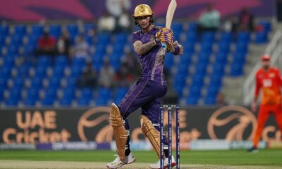 A professional cricket player for the Desert Vipers in mid-swing during a match. The batsman is wearing a dark green and black patterned jersey with red accents, a red helmet, and black protective leg pads. He is holding a wooden cricket bat high in a follow-through motion after playing a shot. The background shows a crowded stadium with purple and blue seating and a "DP World" branded wicket.