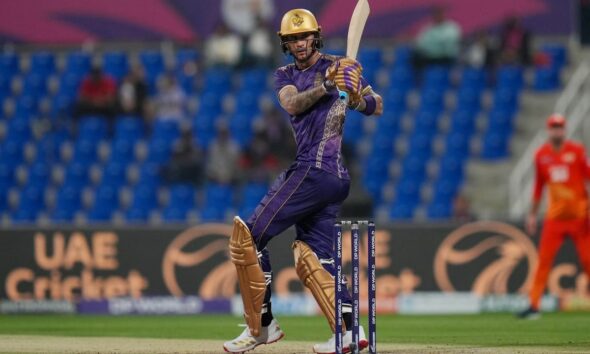 A professional cricket player for the Desert Vipers in mid-swing during a match. The batsman is wearing a dark green and black patterned jersey with red accents, a red helmet, and black protective leg pads. He is holding a wooden cricket bat high in a follow-through motion after playing a shot. The background shows a crowded stadium with purple and blue seating and a "DP World" branded wicket.