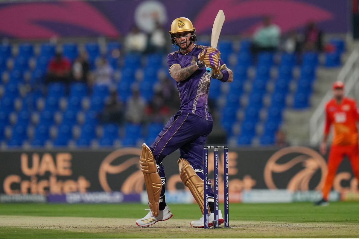 A professional cricket player for the Desert Vipers in mid-swing during a match. The batsman is wearing a dark green and black patterned jersey with red accents, a red helmet, and black protective leg pads. He is holding a wooden cricket bat high in a follow-through motion after playing a shot. The background shows a crowded stadium with purple and blue seating and a "DP World" branded wicket.