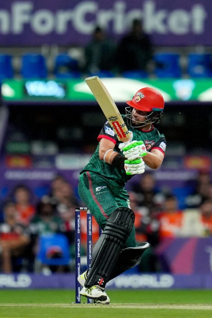 A cricket player from the Abu Dhabi Knight Riders standing at the crease, ready to receive a ball. The player is dressed in a purple and gold uniform with matching gold-colored leg pads and a gold helmet. He holds the bat upward in a standard batting stance. The stadium background features blue seats, a "UAE Cricket" sign, and another player in an orange uniform in the distance.