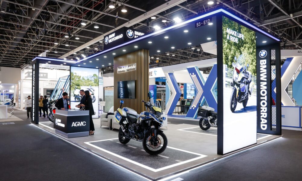 A professional exhibition booth for BMW Motorrad and AGMC at a large indoor trade show. The modern, well-lit stall features a large illuminated pillar with the slogan "MAKE LIFE A RIDE" and a photo of a motorcycle. In the center, a BMW police motorcycle is displayed on a platform designed to look like a road. Staff members in business attire stand behind a branded AGMC counter talking to visitors, while the background shows a high-ceilinged convention hall with other exhibits.