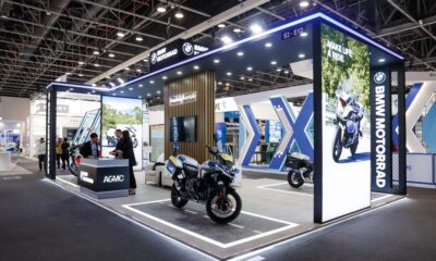 A professional exhibition booth for BMW Motorrad and AGMC at a large indoor trade show. The modern, well-lit stall features a large illuminated pillar with the slogan "MAKE LIFE A RIDE" and a photo of a motorcycle. In the center, a BMW police motorcycle is displayed on a platform designed to look like a road. Staff members in business attire stand behind a branded AGMC counter talking to visitors, while the background shows a high-ceilinged convention hall with other exhibits.