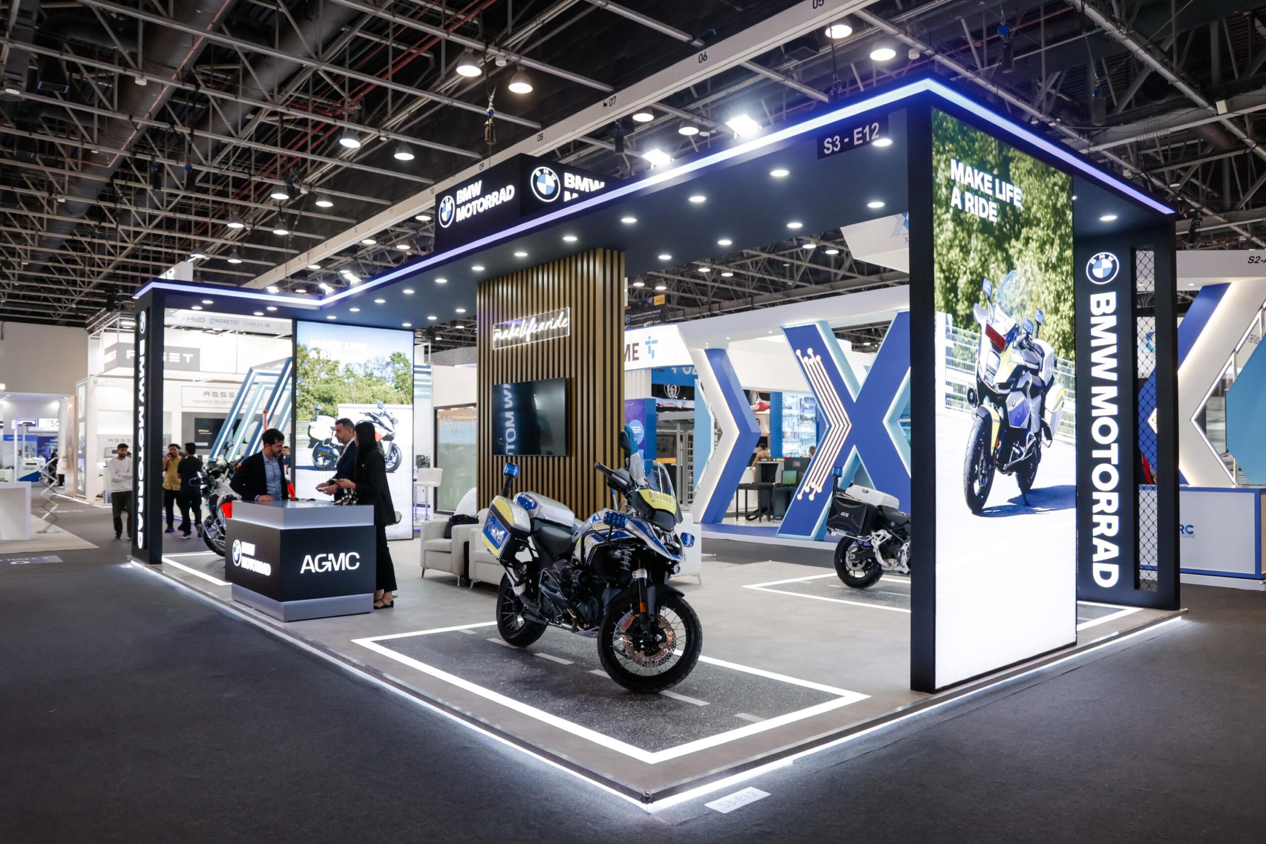 A professional exhibition booth for BMW Motorrad and AGMC at a large indoor trade show. The modern, well-lit stall features a large illuminated pillar with the slogan "MAKE LIFE A RIDE" and a photo of a motorcycle. In the center, a BMW police motorcycle is displayed on a platform designed to look like a road. Staff members in business attire stand behind a branded AGMC counter talking to visitors, while the background shows a high-ceilinged convention hall with other exhibits.