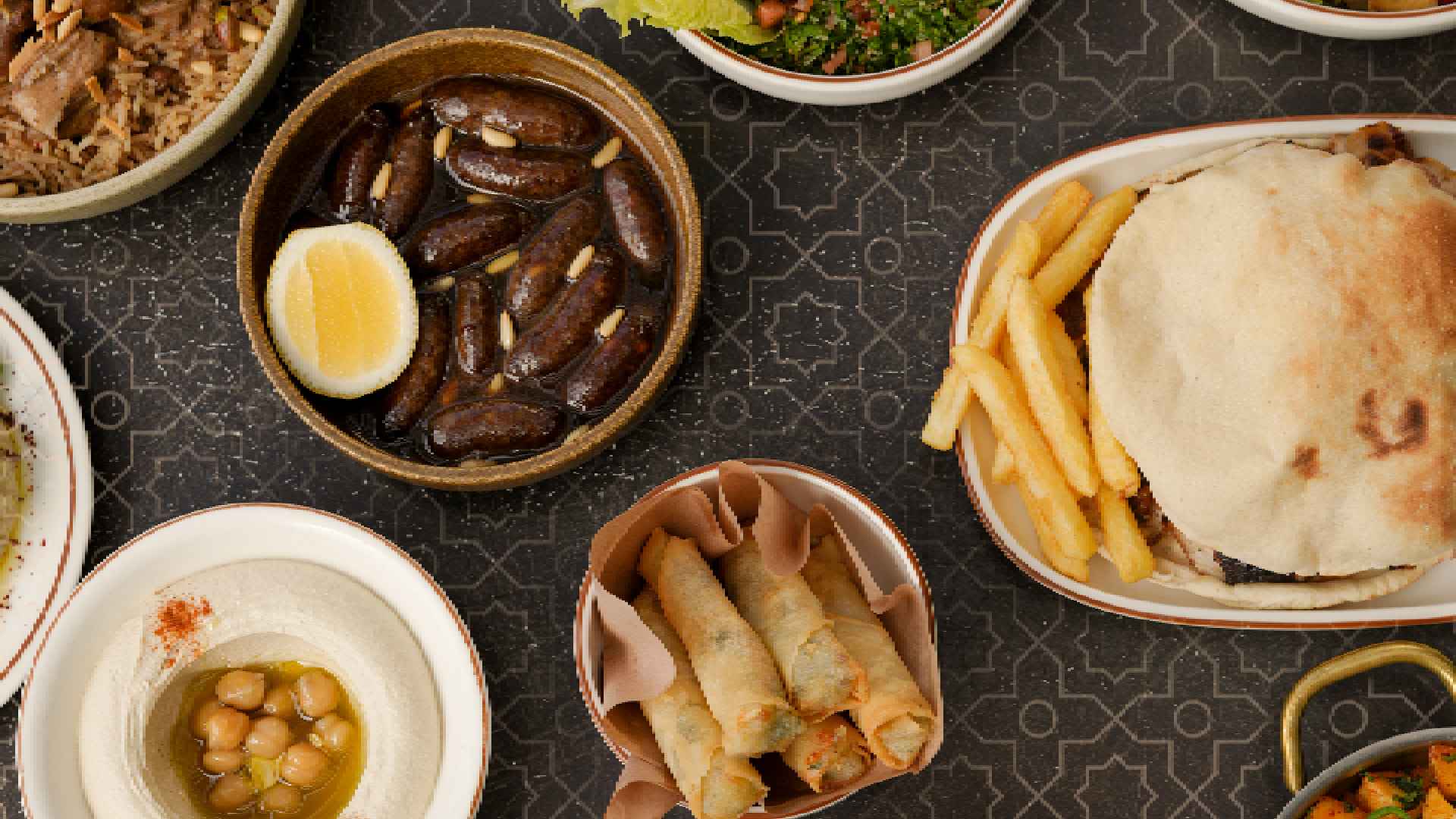 Overhead view of AL HALLAB Lebanese dishes including stuffed vine leaves, hummus with chickpeas, fried spring rolls, a pita sandwich with fries, dates with boiled egg, and mixed appetizers served on patterned table.