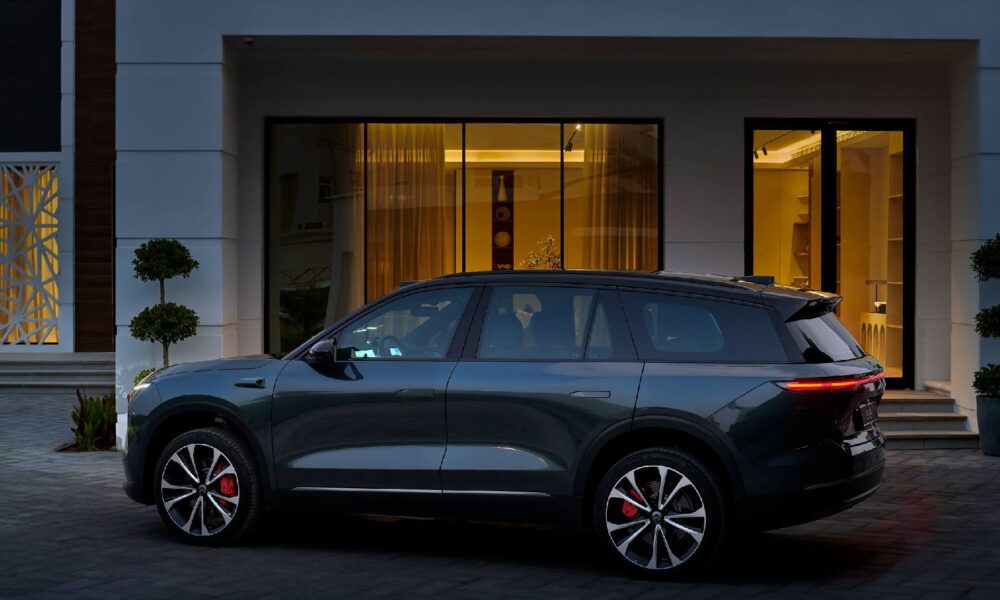 Side view of a dark gray NIO electric SUV parked on a paved driveway in front of a modern house with large glass doors and warm interior lighting.