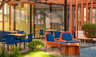 Outdoor dining area at Arcade Eatery & Bar by Aloft featuring blue woven chairs, orange-accent lounge seating, wooden tables, warm table lamps, greenery, and a modern pergola structure with glass walls.