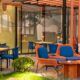 Outdoor dining area at Arcade Eatery & Bar by Aloft featuring blue woven chairs, orange-accent lounge seating, wooden tables, warm table lamps, greenery, and a modern pergola structure with glass walls.