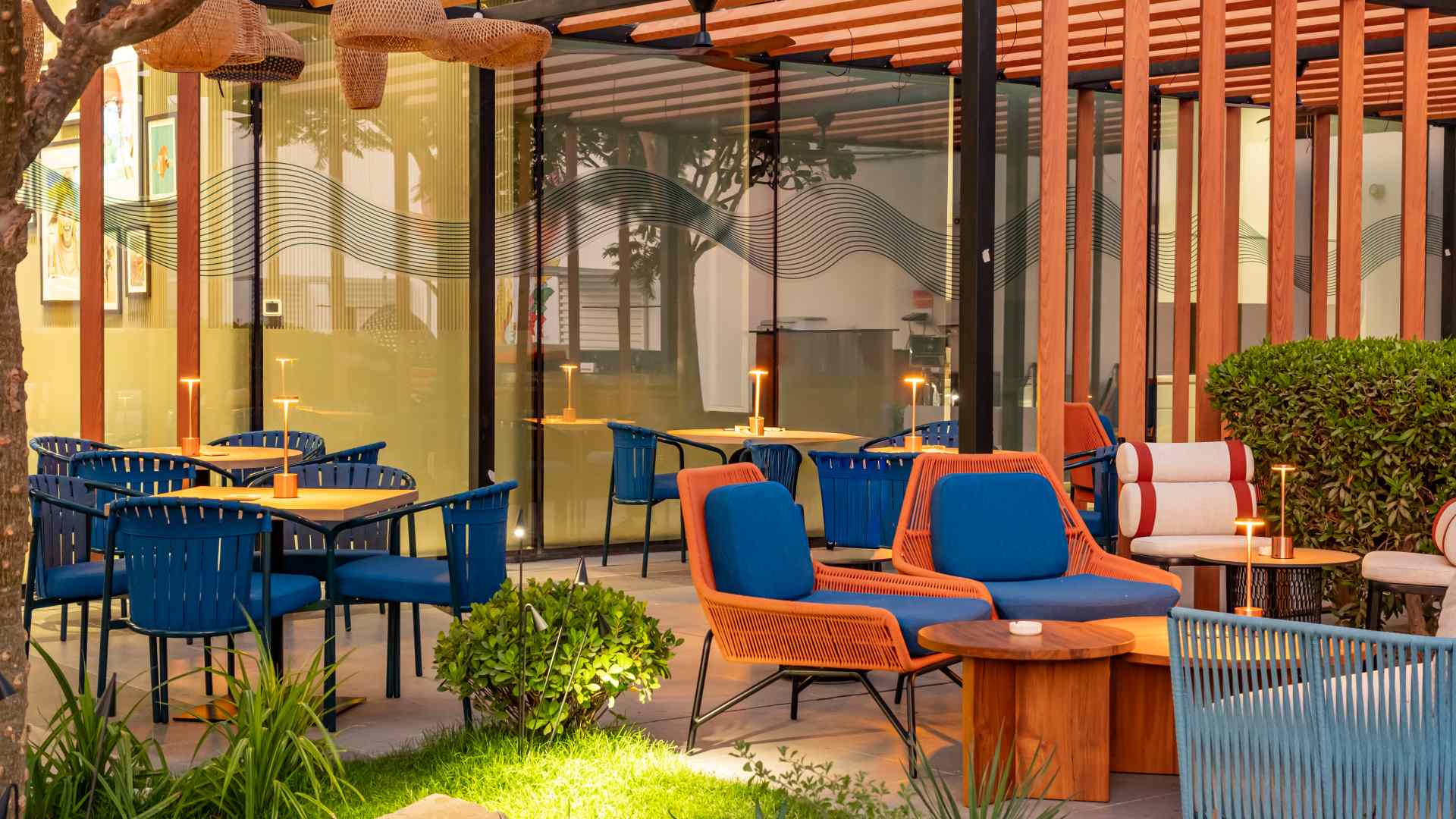 Outdoor dining area at Arcade Eatery & Bar by Aloft featuring blue woven chairs, orange-accent lounge seating, wooden tables, warm table lamps, greenery, and a modern pergola structure with glass walls.