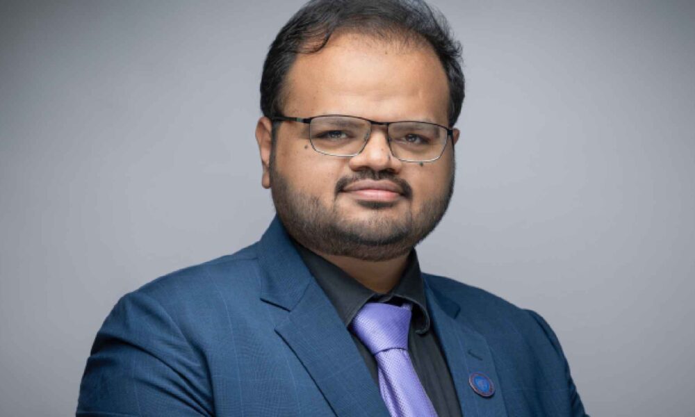 Person wearing a blue suit jacket, black shirt, and light purple tie standing against a plain grey background at Heriot-Watt University Dubai.