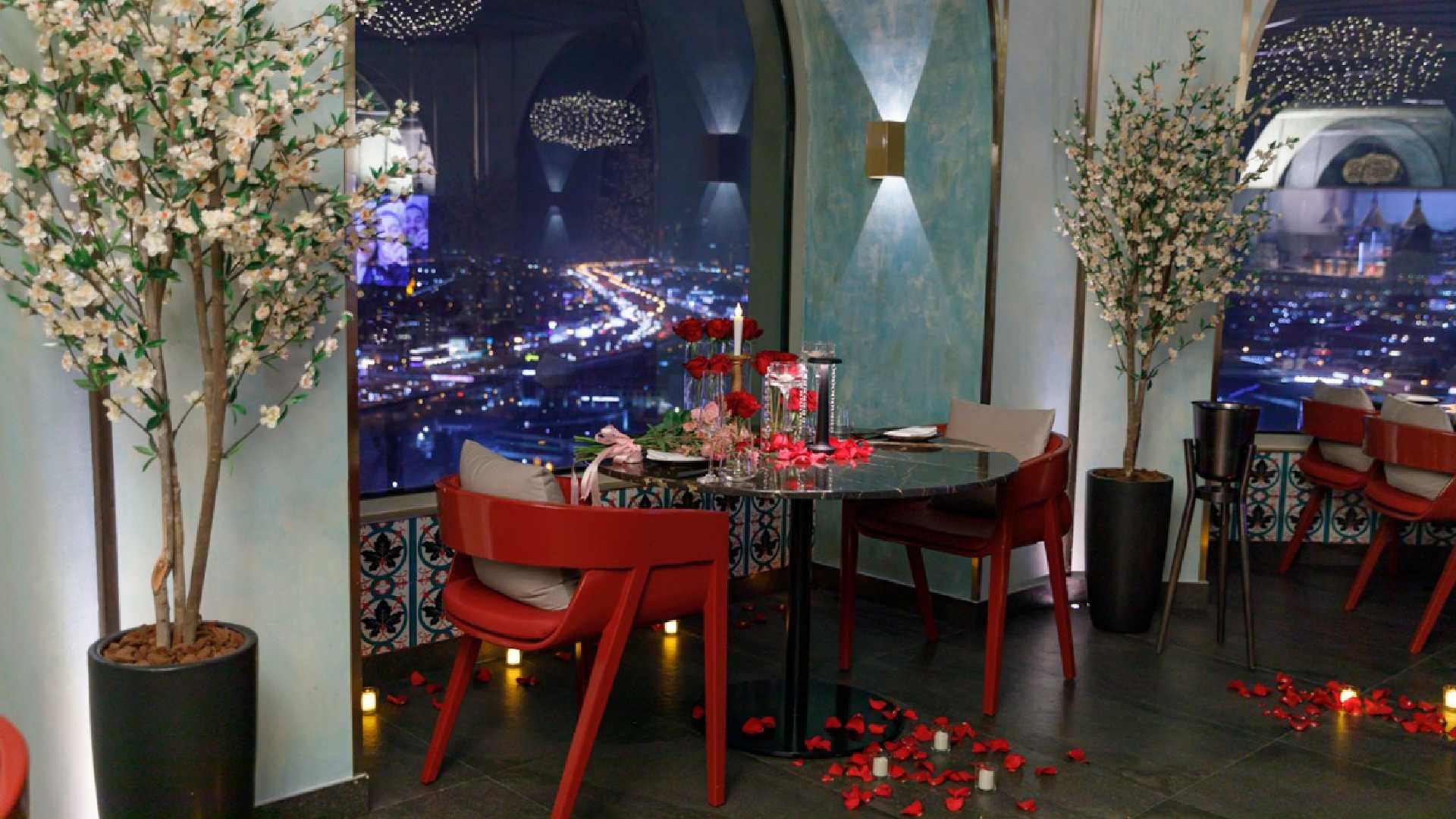 A romantic dining setup at Oanjo featuring a round table decorated with red roses, candles, and rose petals, surrounded by red chairs and floral arrangements, overlooking a vibrant city skyline through large arched windows at night.