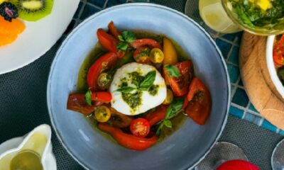 A bowl of burrata served with sliced cherry tomatoes, red and yellow peppers, and basil, drizzled with green pesto, surrounded by additional dishes including a plate of fresh fruit and a drink on a tiled table at Oasis Terrace and Garden.
