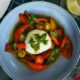 A bowl of burrata served with sliced cherry tomatoes, red and yellow peppers, and basil, drizzled with green pesto, surrounded by additional dishes including a plate of fresh fruit and a drink on a tiled table at Oasis Terrace and Garden.