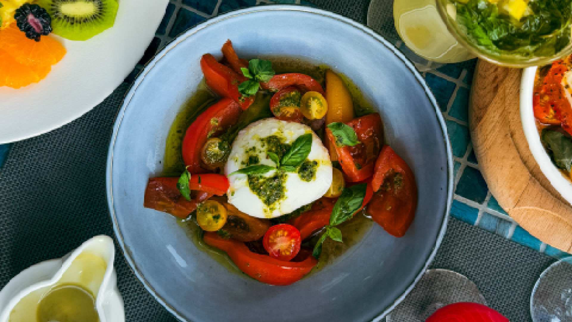 A bowl of burrata served with sliced cherry tomatoes, red and yellow peppers, and basil, drizzled with green pesto, surrounded by additional dishes including a plate of fresh fruit and a drink on a tiled table at Oasis Terrace and Garden.