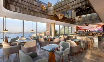 A modern, sunlit restaurant at TUMBI Hotel Dubai The Palm featuring elegant pastel-colored chairs, wooden tables, floor-to-ceiling windows overlooking the waterfront, and a contemporary ceiling design with geometric gold light fixtures and reflective panels.