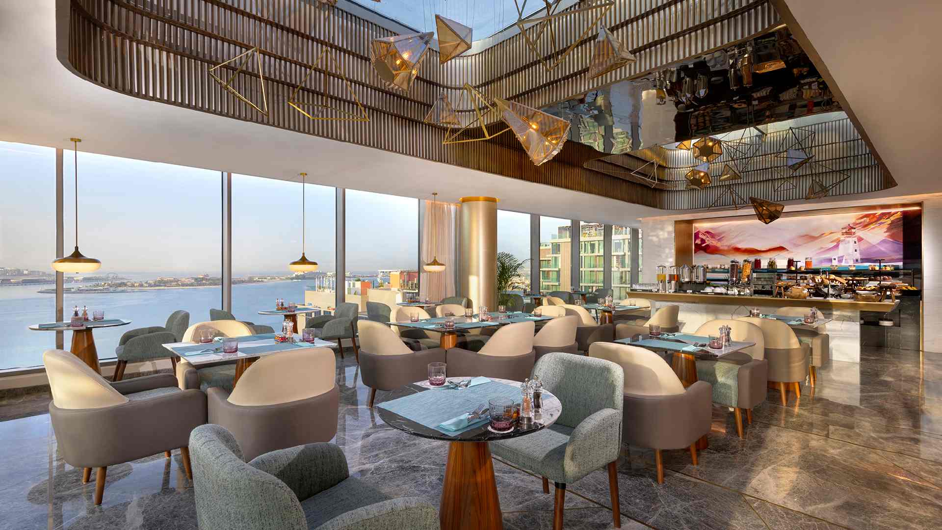 A modern, sunlit restaurant at TUMBI Hotel Dubai The Palm featuring elegant pastel-colored chairs, wooden tables, floor-to-ceiling windows overlooking the waterfront, and a contemporary ceiling design with geometric gold light fixtures and reflective panels.