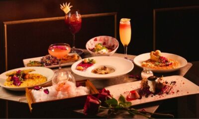 A romantic Valentine’s tasting menu spread at AT THE PERMIT ROOM, featuring multiple plated dishes including risotto, chaat, desserts, and garnished cocktails arranged on a table with rose petals and warm ambient lighting