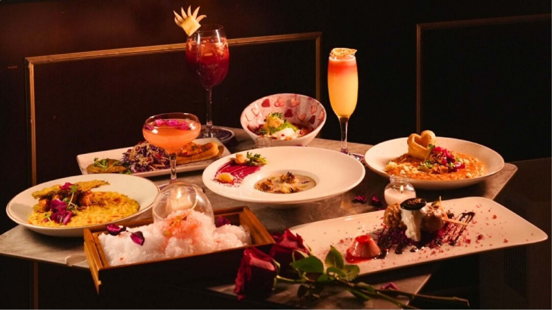 A romantic Valentine’s tasting menu spread at AT THE PERMIT ROOM, featuring multiple plated dishes including risotto, chaat, desserts, and garnished cocktails arranged on a table with rose petals and warm ambient lighting